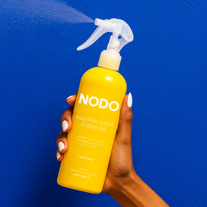 Main vaporisant un flacon NODO Citrus Frais vers un mur bleu, dans une ambiance lumineuse et propre. L’image évoque la fraîcheur instantanée et l’effet purifiant du neutralisant d’odeurs.