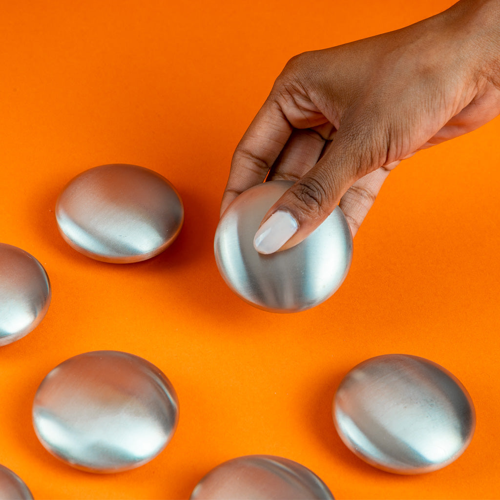 Main tenant un savon en inox NODO sur fond orange vif – savon anti-odeurs naturel qui élimine les odeurs de poisson, d’oignon, d’ail et de soufre laissées sur les mains après la cuisine.